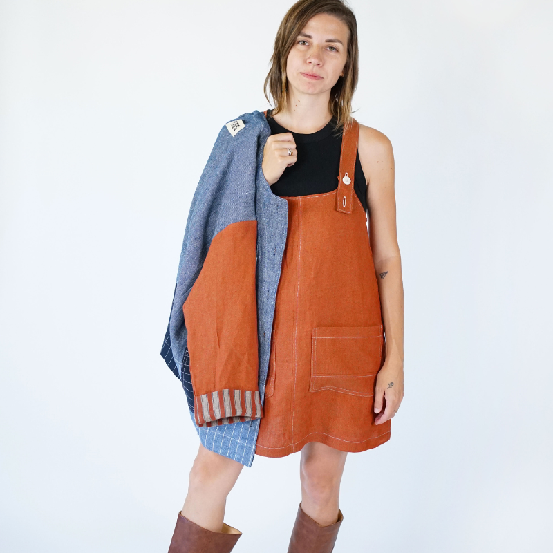 Woman wearing a brown leather pinafore dress with a blue plaid shirt over it on a white background