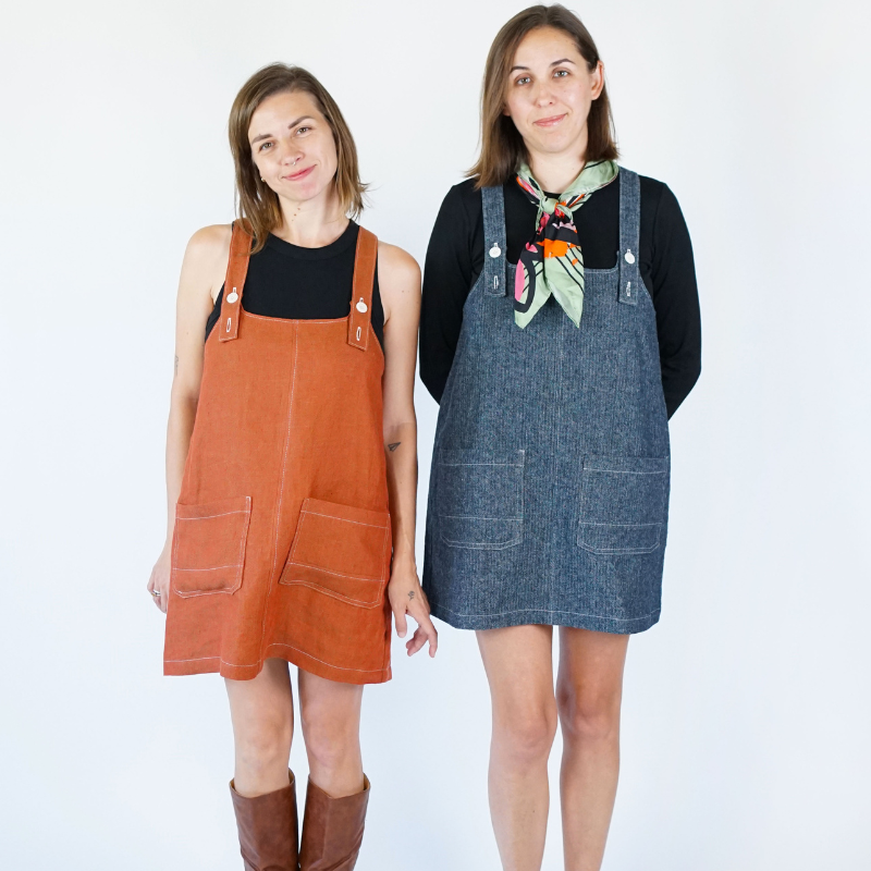 Two women wearing orange and blue denim pinafore dresses on a white background