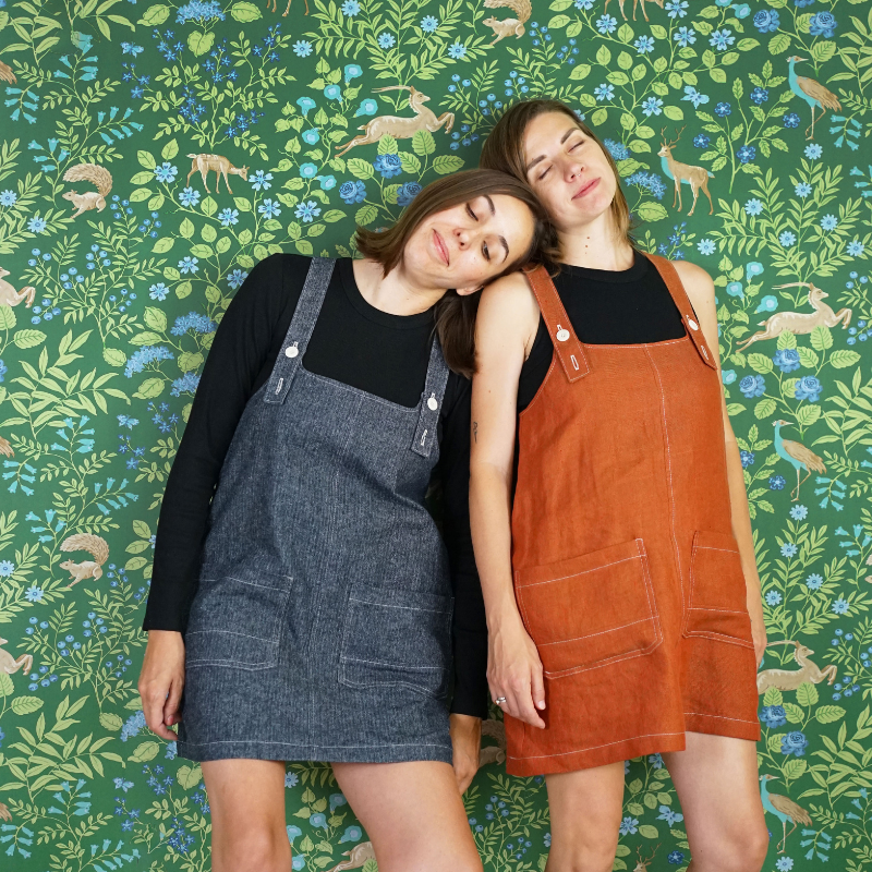 Two women wearing denim and brown pinafore dresses against a green floral patterned wall.
