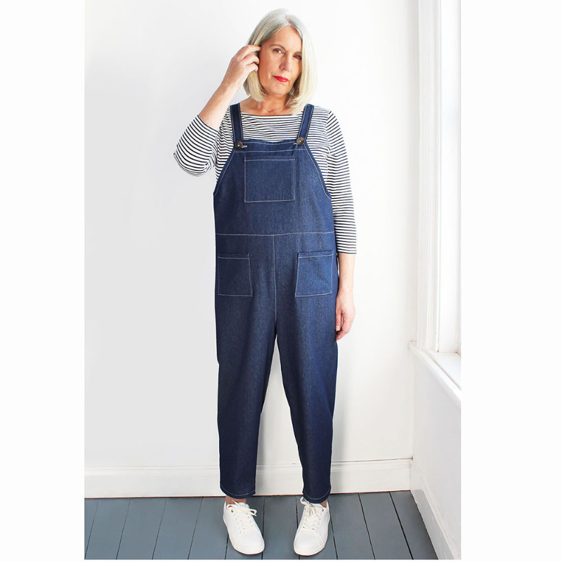 Person wearing blue denim overalls with a striped shirt underneath, standing against a white wall.
