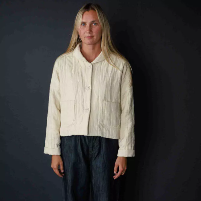 A person standing against a dark background, wearing an oversized beige jacket with a rolled collar, buttoned front, and patch pockets.