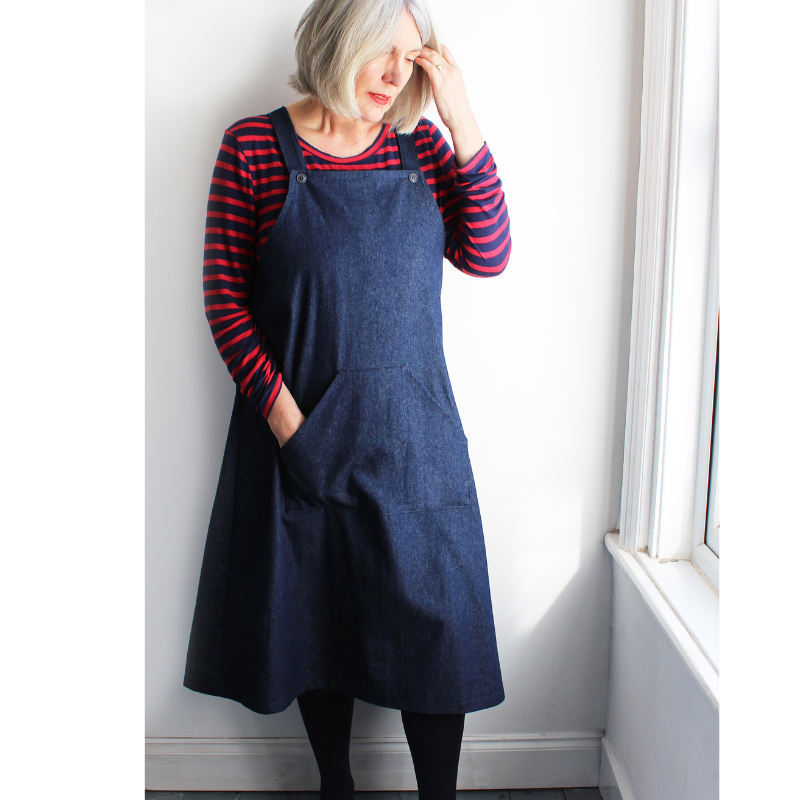 Woman wearing a blue denim pinafore dress over a red and black striped shirt against a white wall.