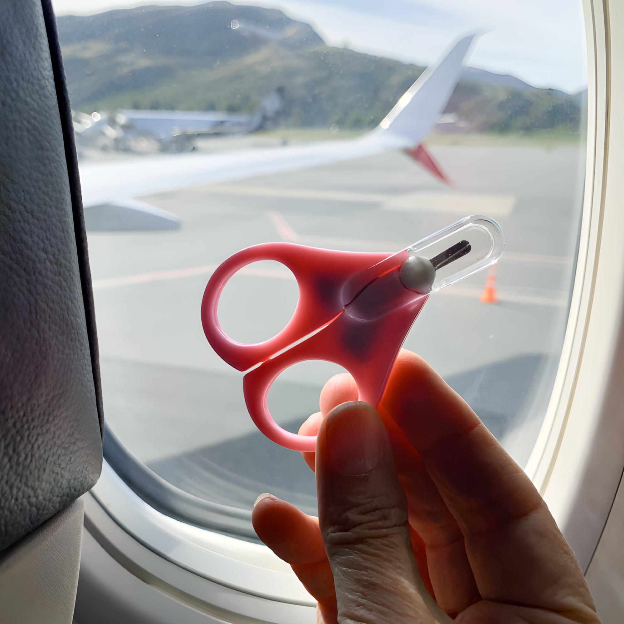 Pink travel scissors held in front of an airplane window with a view of the tarmac.