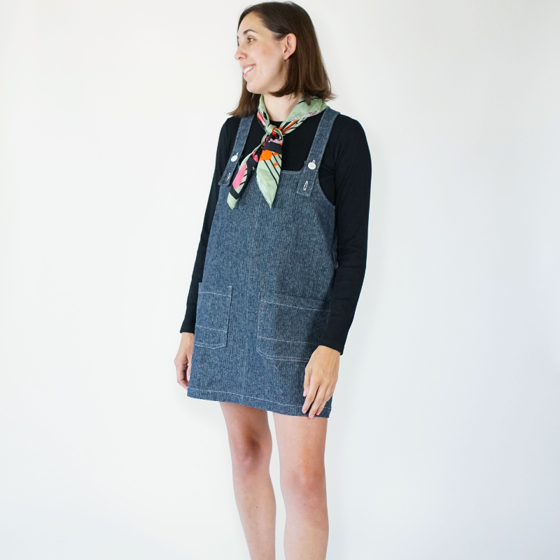 Woman wearing a denim pinafore dress over a black top with a colorful scarf on a white background
