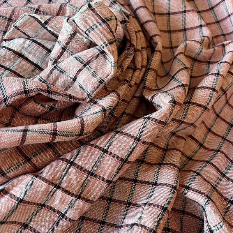 A close-up of draped plaid fabric with a dusty pink base and thin black and green intersecting lines forming a check pattern.