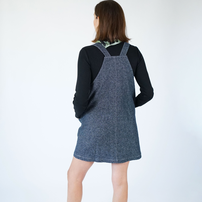 Person wearing a denim pinafore dress over a black top on a white background