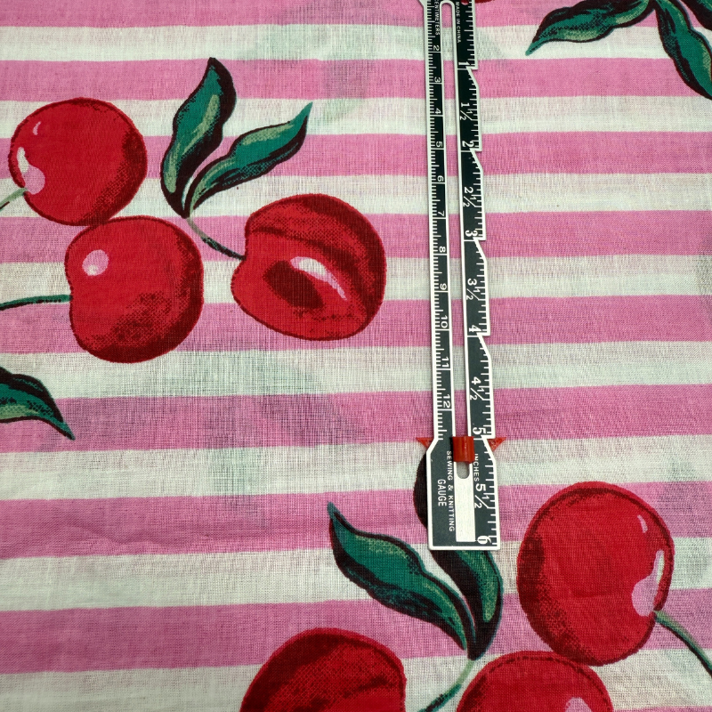 Fabric with red cherries and green leaves on a pink and white striped background, with a ruler for scale.