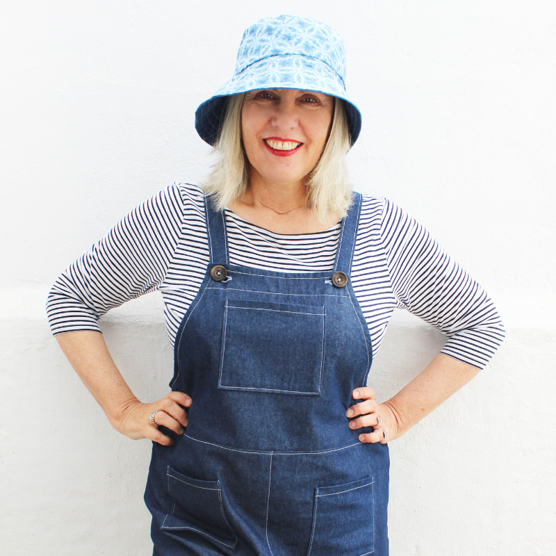 Woman wearing a blue denim overall dress with a striped shirt underneath, against a plain background.